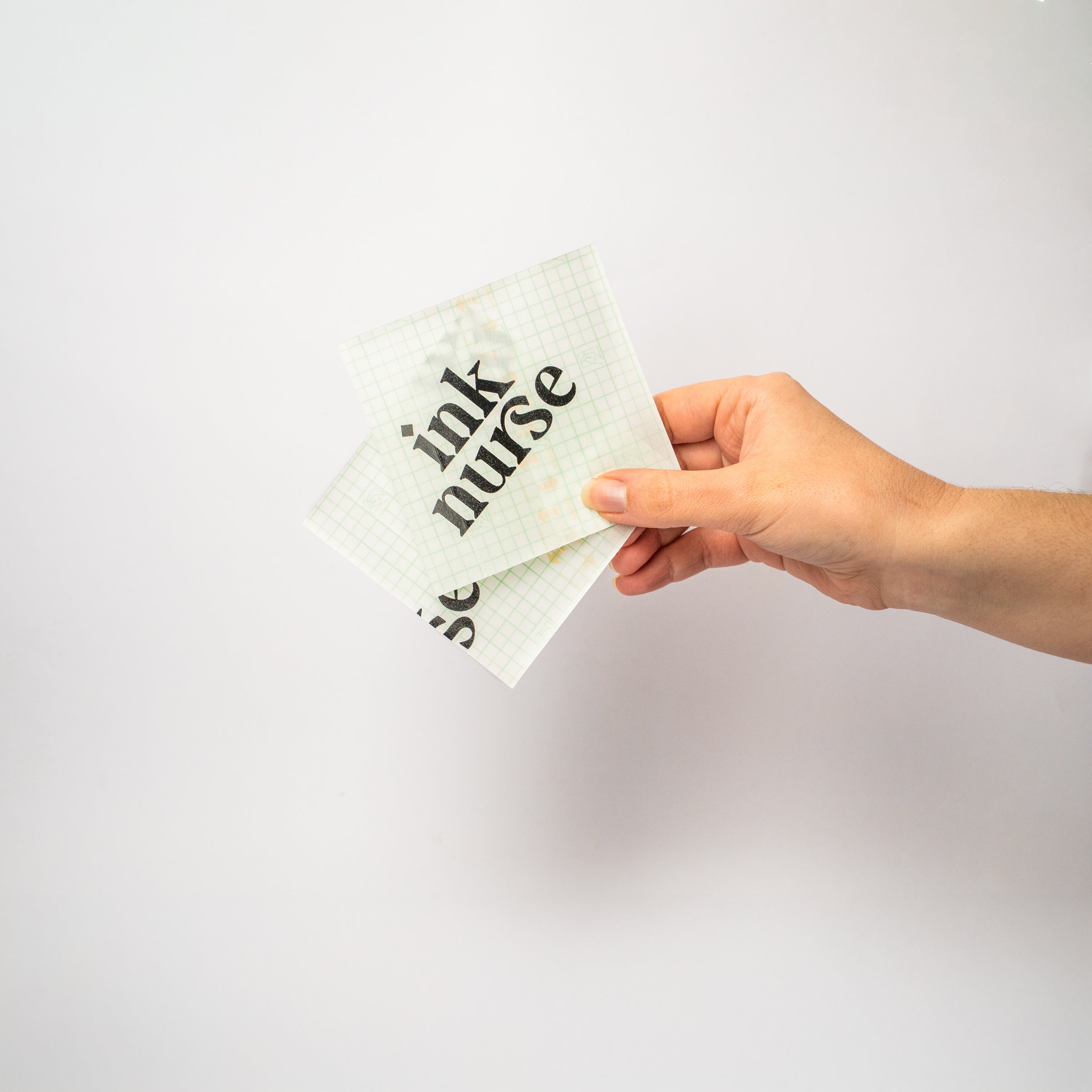 Hand holding 2 squares of second skin with 'ink nurse' logo printed on the tattoo film on a light gray background